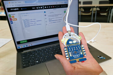Eine Hand hält vor einem Laptop eine per Kabel verbundene Platine in Form eines Roboters mit leuchtenden LED.