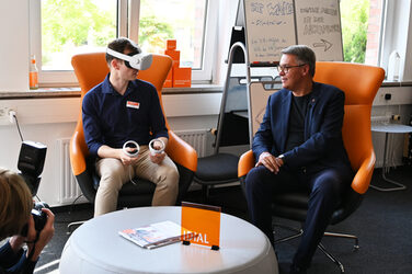 Jonas Sorgalla und OB Thomas Westphal unterhalten sich in Ohrensesseln sitzen über eine VR-Anwendung. Jonas trägt eine VR-Brille.