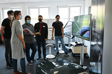 A group of people stand in front of a Turfrob, behind them a television with videos about the Turfrob and a roll-up about the Turfrob.