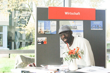 Another exhibitor table of the "Marketplace of Opportunities": a young man sits at the booth and smiles into the camera__ Another exhibitor table of the "Marketplace of Opportunities": a young man sits at the booth and smiles into the camera