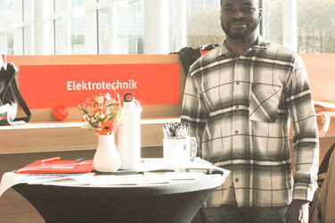 Ein weiterer Ausstellertisch des "Marktplatzes der Möglichkeiten": Ein junger Mann steht, in die Kamera lächelnd, an einem Messestand__Another exhibitor table of the "Marketplace of Opportunities": A young man stands at a booth, smiling into the camera