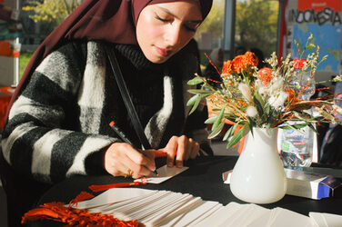 Eine junge muslimische Frau schreibt im Sonnenlicht an einem Tisch eines Messestands__A young muslim woman writes in the sunlight at a table of a booth