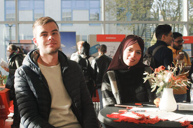 A young Muslim woman and a young man smile into the camera in the sunlight at a table of a booth, at the "Marketplace of Opportunities"__ A young Muslim woman and a young man smile into the camera in the sunlight at a table of a booth, at the "Marketplace of Opportunities"