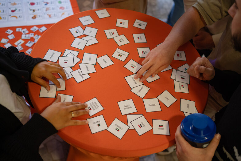 Hände die an einem organgefarbenen Stehtisch ein Wortspiel mit Karten spielen.__Hands playing a word game with cards at an orange bar table.
