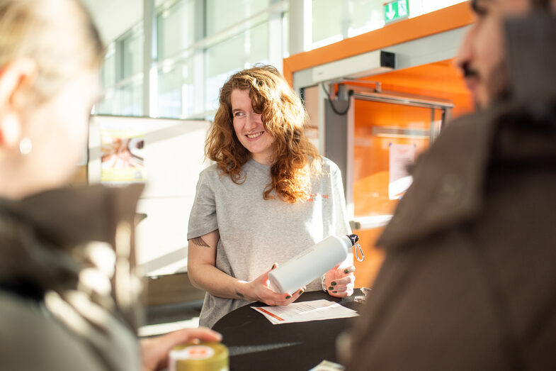 Eine junge rothaarige Frau hält eine Trinkflasche der Fachhochschule Dortmund in den Händen und lächelt im Gespräch.__A young red-haired woman holds a water bottle from Dortmund University of Applied Sciences and Arts in her hands and smiles in conversation.