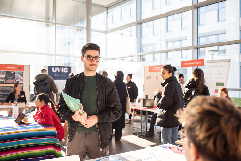 Ein junger Mann mit einer Mappe unter dem Arm steht an einem Messestand.__A young man with a folder under his arm stands at a trade fair stand.