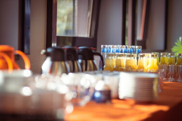 Kaffekannen, Gläser, Teller, Tassen und Orangensaft auf einer orangen Tischdecke im Sonnelicht an einem Buffet__Coffee pots, glasses, plates, cups and orange juice on an orange tablecloth in the sunlight at a buffet