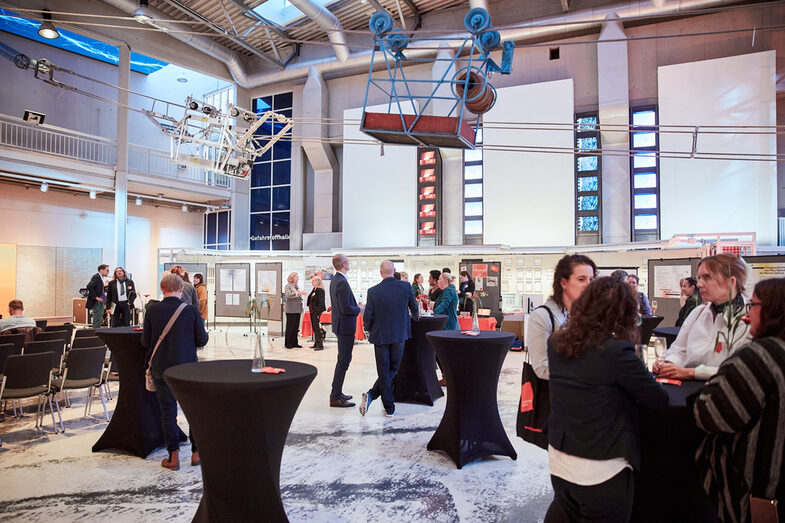 Ein Panoramablick in die Veranstaltungshalle, mit zahlreichen Besuchern in Gesprächen an Stehtischen__ A panorama view into the venue, with visitors standing at bar tables in conversation