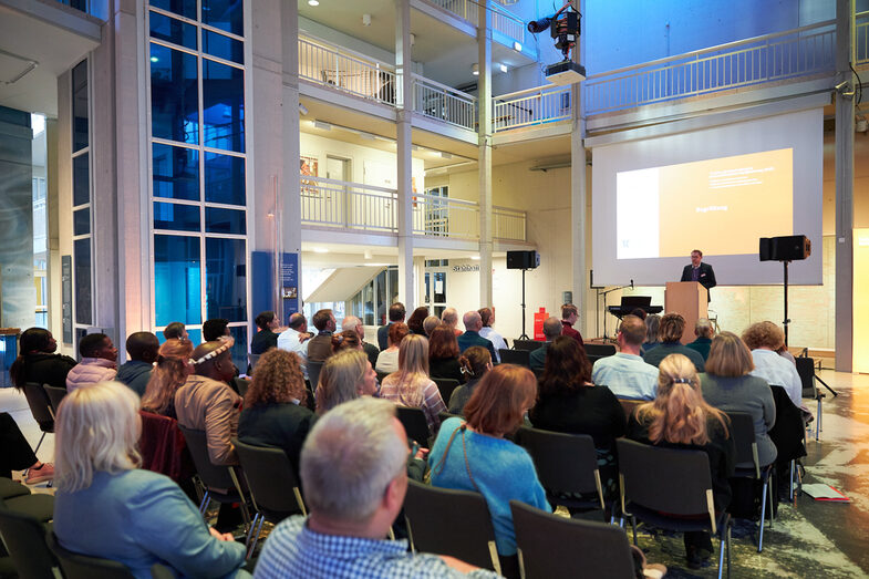 Ein Blick über die besetzten Stuhlreihen auf die Bühne beim Eingangsvortrag, mit einem Sprecher am Rednerpult__A view across the rows of taken chairs during the introductory presentation of the event, with a speaker at the speaker's desk