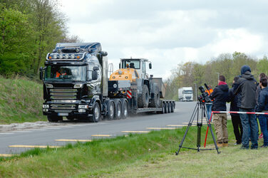 Photo of a 40-ton truck with a tipper loader on the loading area. Several people are standing at the side of the road, a camera is set up.