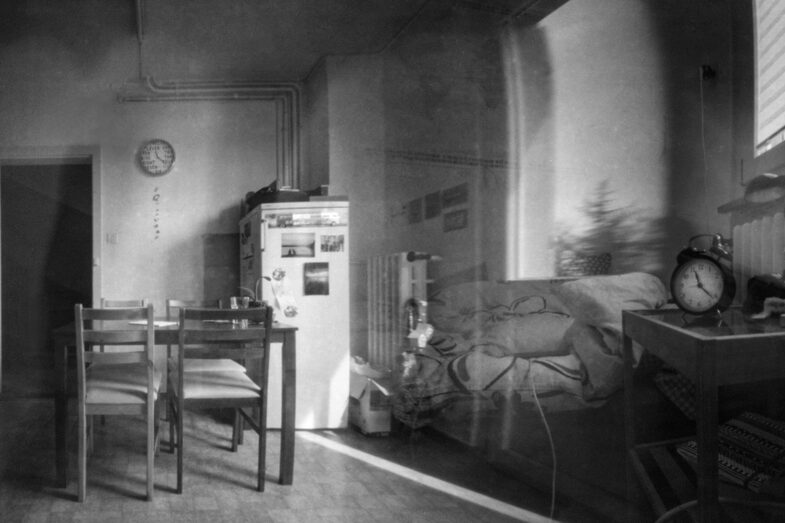 Schwarz-Weiß-Foto, das aussieht, als sei es durch eine Scheibe hindurch aufgenommen worden. Zu sehen ist ein Zimmer mit Tisch und Stühlen, Kühlschrank, Bett und weiteren Möbeln. Keine Menschen. Die Wanduhr zeigt 11.20 Uhr.