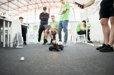 Ein Roboter mit Greifarm steht auf dem Boden, vor ihm liegt eine kleine Kugel. Im Hintergrund sind Personen unscharf zu sehen.