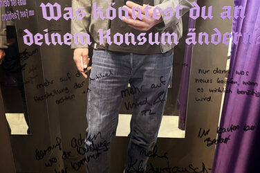 A person stands in front of a mirror and takes a photo of themselves with their cell phone; the person can only be seen in the mirror, their head is beyond the edge of the picture. The following is written in purple on the mirror: "What could you change about your consumption?" Below this are several comments left in different handwriting in black felt-tip pen, for example "buy more second-hand".