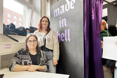 Two people are sitting or standing in the left half of the picture and looking into the camera. To their right is a black changing room with the inscription "made in hell".