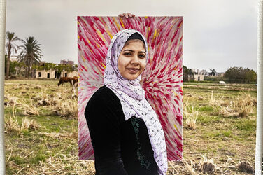 A person with a light headscarf and dark top is standing in front of a colorful, abstract painting that someone is holding up right behind her. Around it is a meadow where cattle are grazing.