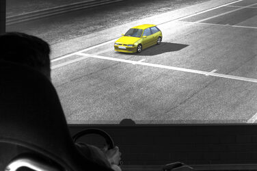 Auf einem großen Bildschirm ist ein gelbes Auto zu sehen. Schemenhaft ist im Vordergrund eine Persona an einem Lenkrad zu sehen.