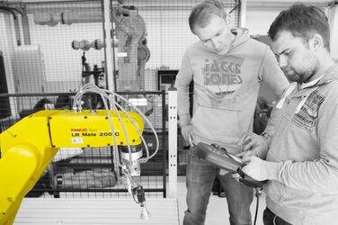 Zwei junge Männer stehen im Robotik-Labor vor einem Industrieroboter und steuern die Maschine.