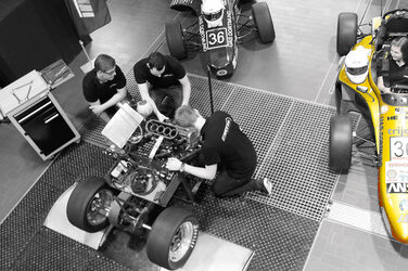 A bird's eye view of a yellow racing car. Next to it, students are working on an engine.