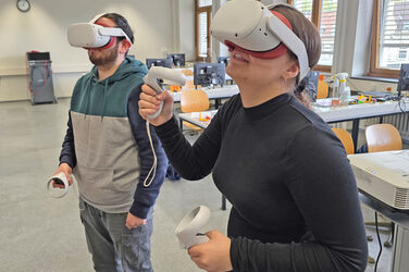 Two people are standing in a seminar room, each wearing VR goggles and holding control elements in their hands.