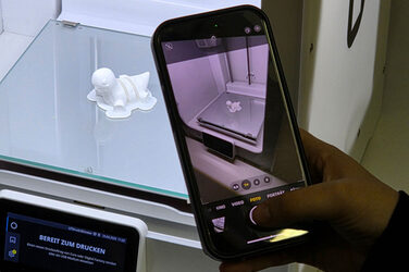 A hand holds a smartphone to take a photo of a miniature figure lying in a 3D printer.