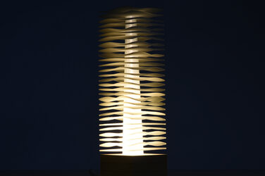 Nachtaufnahme einer säulenartigen, erleuchtenden, skulpturalen Leuchte auf einem Holzsockel