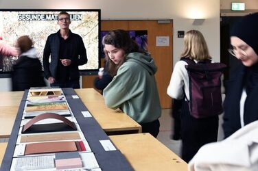 Im Vordergund sind einge Studeierende bei der Metrachtugn von Materielmustern zu sehen, während Daniel Horn die Ausstellung im Rahmen einer Vernissage eröffnet