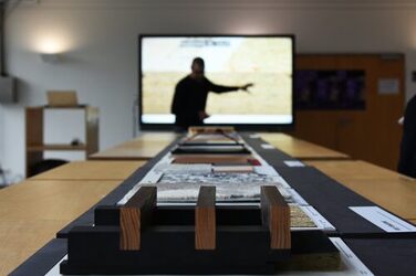 Im Vordergund sind unterscheildiche Materialien aus der Ausstellung zu sehen, während im Hintergrund, unscharf dargestellt, der Gastreferent seinen Vortrag vor einem Monitor hält