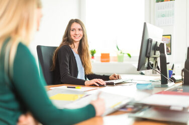 Foto einer jungen Frau an einem Arbeitsplatz mit Rechner. Sie schaut zu einer weiteren Frau am Schreibtisch, die Notizen macht - diese ist nur zum Teil zu erkennen.