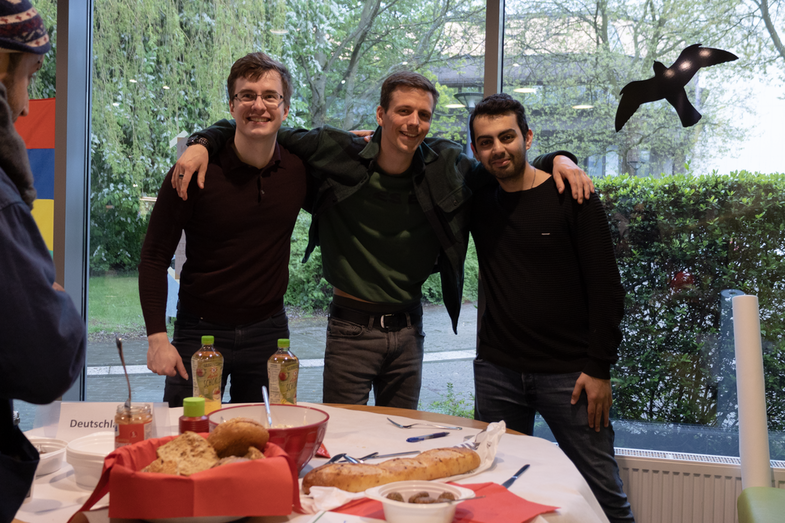 A cultural stand from Germany can be seen. There is bread, sausages and apple spritzer on the table. Behind the table are three students looking after the stand.