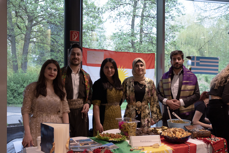 A cultural stand from Kurdistan is on display. On the table is a rich buffet with various dishes and drinks typical of the country. Behind it is a group of five students running the stand. In the background is a Kurdish flag.