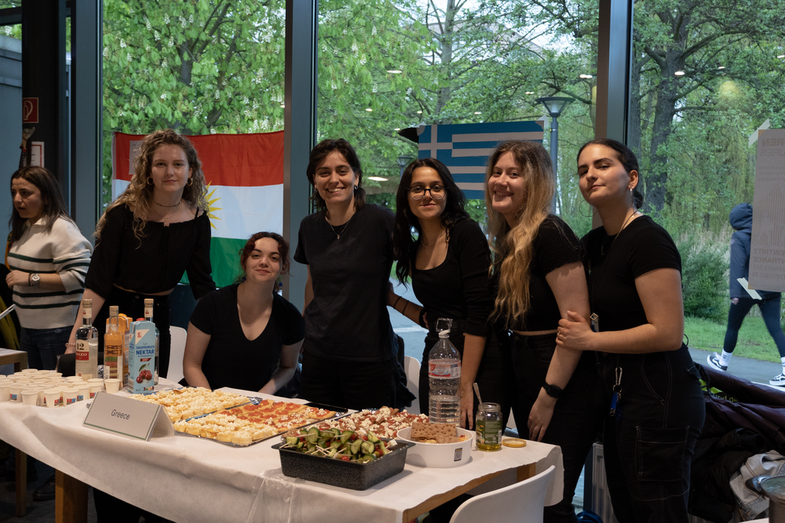 A cultural stand from Greece can be seen. On the table is a rich buffet with various dishes and drinks typical of the country. Behind it is a group of six students who are in charge of the stand. In the background is a Greek flag.