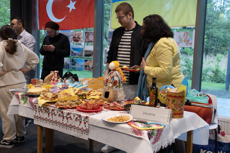 A cultural stand of the Ukraine can be seen. On the table is a rich buffet with various dishes typical of the country and colorful tablecloths. Behind it are two students who are in charge of the stand. In the background is a Ukrainian flag.