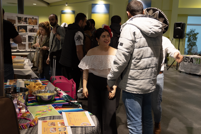 In the background is a group of students. On the left in the foreground is a cultural stand from Mexico. To the right is the student in charge of the stand, talking to visitors.