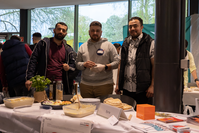 A cultural stand from Syria is on display. On the table is a rich buffet with various dishes and drinks typical of the country. Behind it is a group of three students who are in charge of the stand.