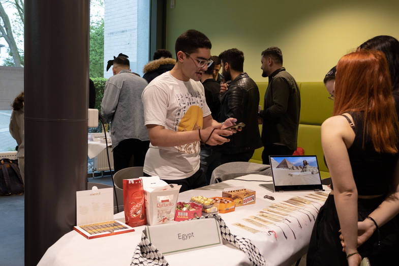 A cultural stand from Egypt can be seen. On the table are sweets and a laptop on which pictures of pyramids can be seen. Behind it, a student can be seen talking to several visitors.
