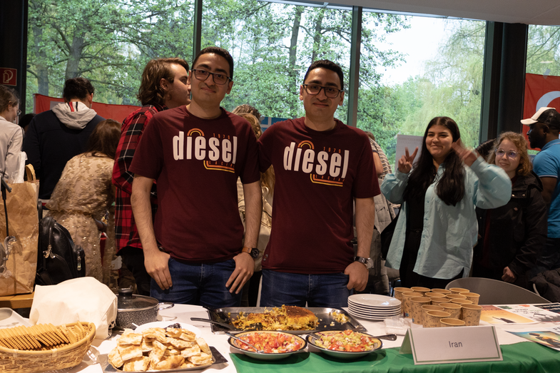 A cultural stand from Iran is on display. On the table is a rich buffet with various typical Iranian dishes and drinks. Behind it are two students who are in charge of the stand.