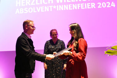 Vice Dean Ralf Dietz congratulates graduates on completing their studies, with Chancellor Svenja Stepper in the background in front of the colorful illuminated screen.