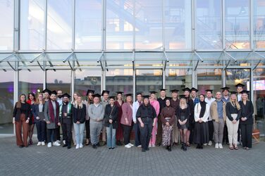 Group photo of the graduates with graduation hats in front of the faculty's glass facade.