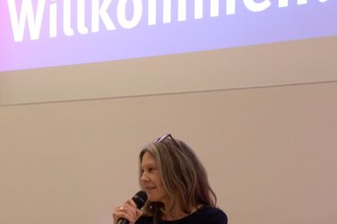 Dean Prof. Diana Reichle at the lectern on the stage of the lecture hall, in the background a slide projected onto the screen welcomes the guests.