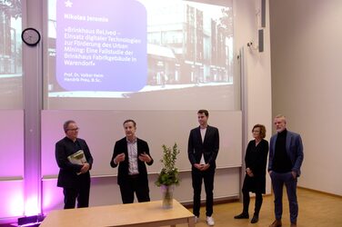 Lecturers with a student on the stage of the lecture hall during the award ceremony; a visualization of the design is projected onto the screen in the background.