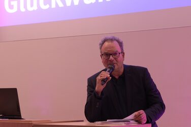 Mr. Otto Weber leans against the lectern on the stage of the lecture hall, which is illuminated in purple.