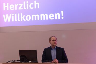 Prof. Dr. Franz Vogler at the lectern on the stage of the lecture hall, which is illuminated in purple.