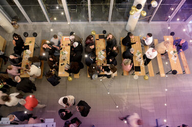 Die Gäste sitzen und stehen an langen Holztischen im festlich geschmückten Foyer des Fachbereichs mit Lichterketten und Ballons. Die Atmosphäre wirkt gesellig und entspannt.