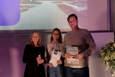 ChatGPT:

Die Dekanin und zwei Studierende stehen auf der Bühne und lächeln in die Kamera, zwei von ihnen halten Urkunden in den Händen. Im Hintergrund ist eine Präsentationsfolie und violette Beleuchtung zu sehen.