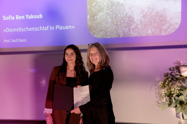 Die Dekanin gratuliert einer Studentin auf der Bühne, sie halten gemeinsam eine Urkunde, während im Hintergrund eine Präsentationsfolie mit Namen und Projekttitel zu sehen ist. Die Szene ist in violettes Licht getaucht.