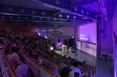 Der gefüllter Hörsaal, in dem eine Frau vorne spricht. Die Projektion zeigt „Herzlich Willkommen!“ in lila Beleuchtung.