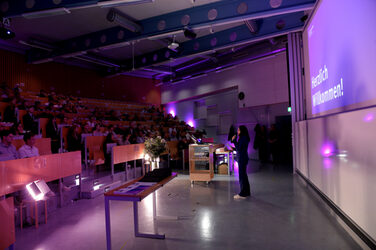 Moderatorin Eva Paar im Hörsaal vor einem großen Publikum. Die Beleuchtung ist in violettem Licht gehalten, und auf der Leinwand steht „Herzlich Willkommen!“.
