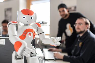 Photo of NAO robot Eva standing on the table in front, arms raised. In the background two men, one at the computer, both looking at the robot. __ <br>NAO robot Eva stands in front of the table, arms raised. In the background someone is sitting at a laptop and talking to the person standing next to it. Both look at the robot.