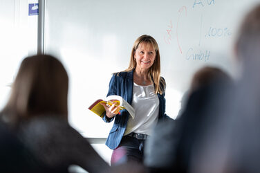 Eine Professorin steht lächelnd vorne im Raum vor einem weißen Board mit Notizen. In ihrer Hand hält sie ein Buch und schaut zu ihren Studierenden, die im Raum sitzen. Von den Studierenden sind nur ein paar unscharf von hinten erkennbar. __ <br>A female professor stands in front of the room smiling in front of a white board with notes. She holds a book in her hand and looks at her students who are sitting in the room. Only a few of the students are out of focus from behind.