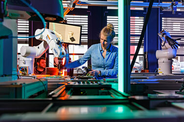 Foto von einem Förderband mit Roboterarmen, hinter dem eine Mitarbeiterin steht, die mit der rechten Hand zu einem Roboterarm greift. __ Behind a conveyor belt with robot arms stands an employee who reaches for a robot arm with her right hand.